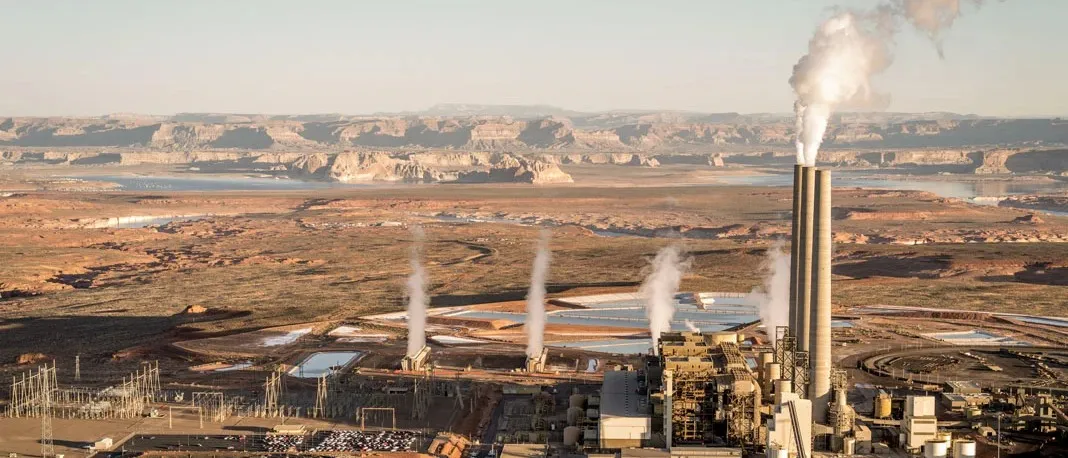 Aerial view of a desert power generating station with tall stacks and steam plumes, evaporation ponds beside the plant, and a lake in the distance